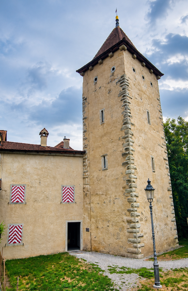 Stone medieval tower attached to a beige building with red-tiled roof and chevron-patterned shutters, open doorway, and a lamp post nearby.