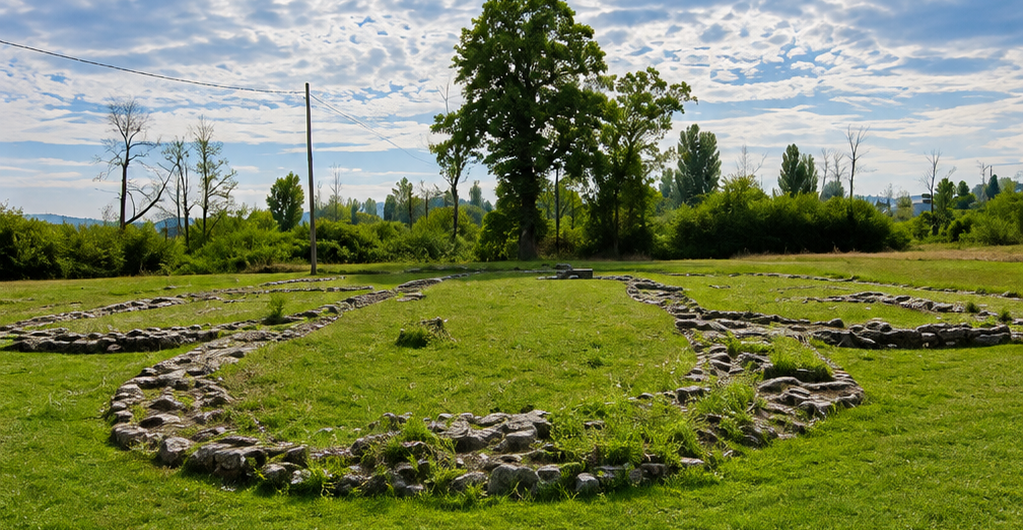 Circular stone-wall ruins arranged in concentric rings on a green field, with trees and a blue, partly cloudy sky in the background.
