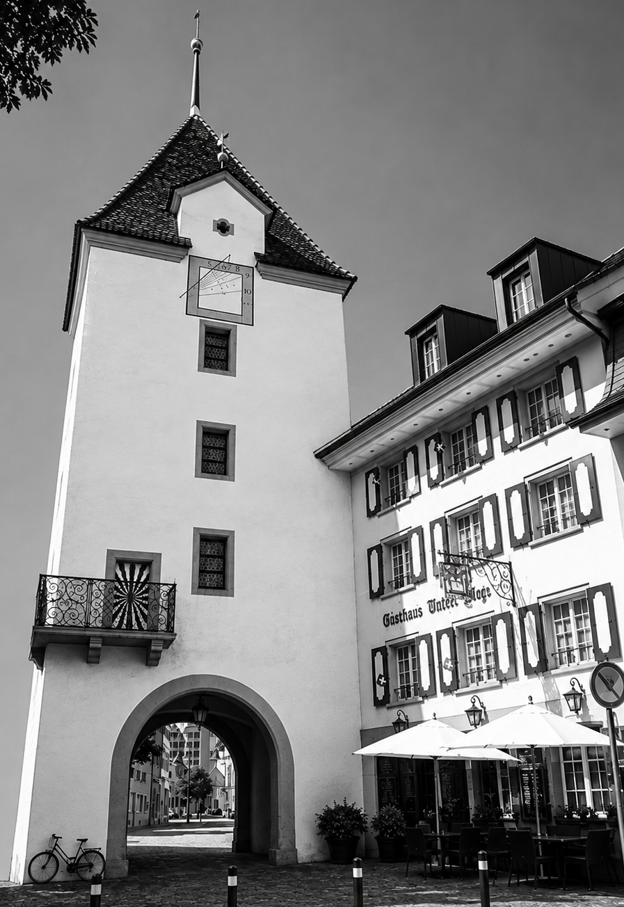 Historic European street scene with a tall clock tower and arched gateway leading to a cobblestone street. Café umbrellas and outdoor seating along the building. Black-and-white photo.