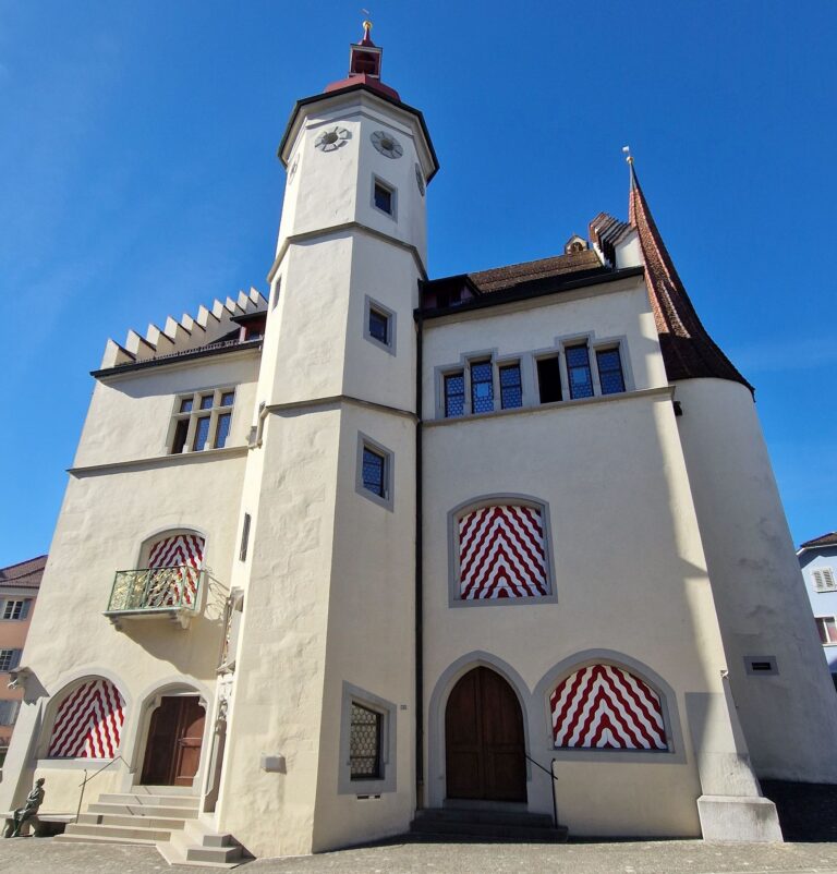 White building with a tall tower, red roof accents, and red-and-white striped window coverings set against a clear blue sky and sunlight on the facade.