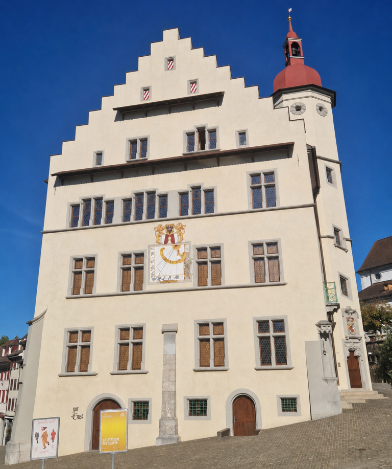 Facade of a historic beige building with stepped gables and a red-domed tower, set in a cobblestone square under a blue sky.